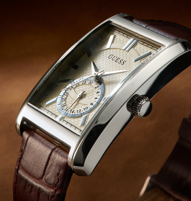 A close-up of a Guess wristwatch with a silver-tone rectangular case, cream textured dial, blue outer ring around the subdial, and a brown leather strap against a warm brown gradient background.