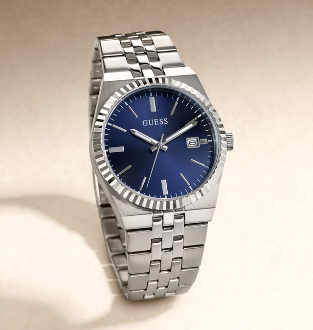 A Guess men's stainless steel wristwatch with a blue dial, silver-tone fluted bezel, and matching steel link bracelet, photographed on a warm beige studio background.
