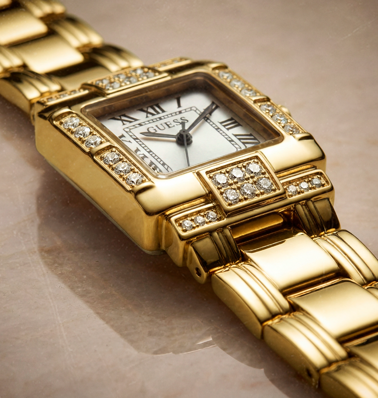 A close-up shot of a gold-tone GUESS watch with a diamond-encrusted bezel and a white dial, resting on a neutral beige surface.