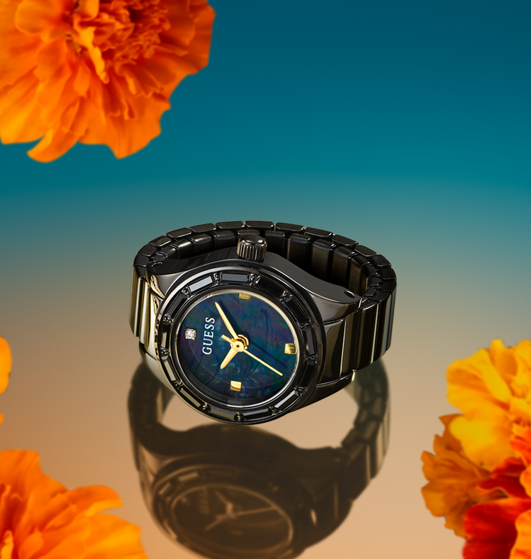A GUESS ring watch with a black ceramic and stainless steel link bracelet, a black bezel, and a dark blue dial with gold accents, presented on a reflective surface with orange flowers against a blue gradient background.