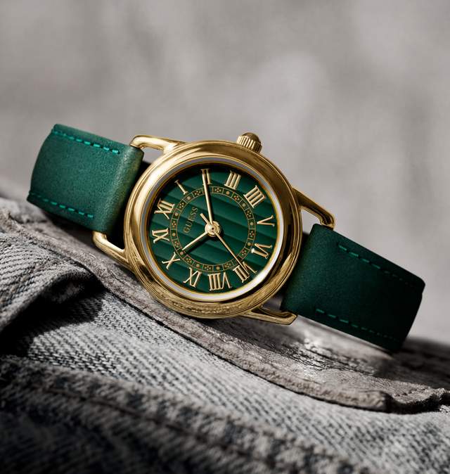A vintage-style wristwatch with a green dial, gold-tone case, and green leather strap, resting on a textured gray fabric surface against a soft gray background.