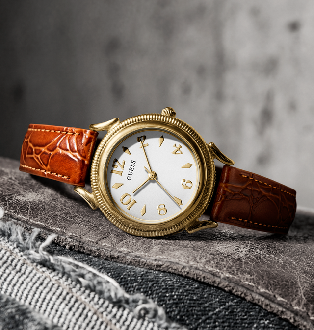 A Guess wristwatch with a gold-tone round case, white dial, and brown leather strap, resting on a textured gray surface.