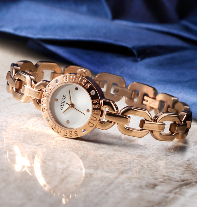 Rose gold chain-link watch with 'Guess' branding on a reflective surface. GW1019L4