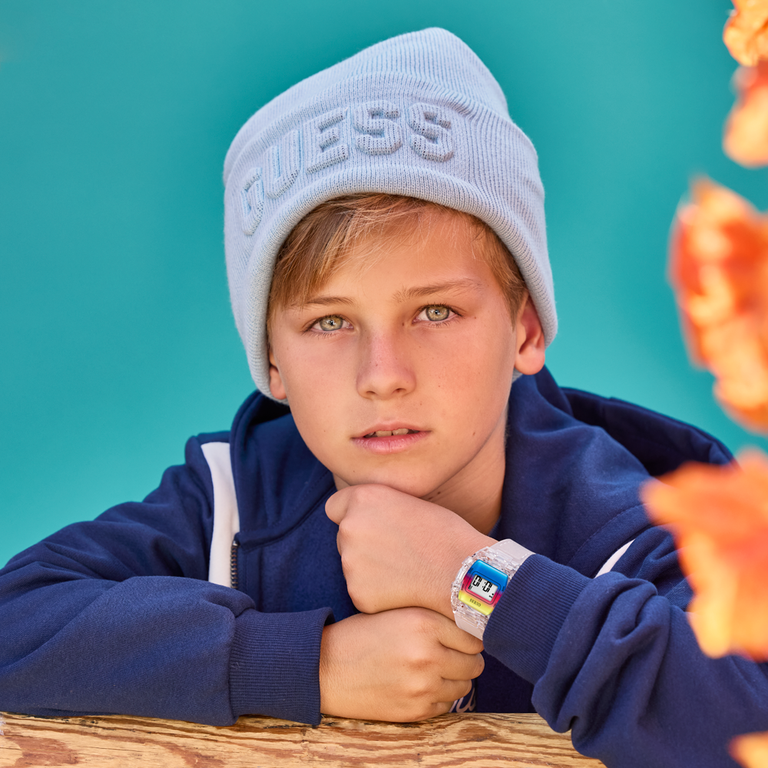 Child wearing a blue hoodie and light blue 'Guess' beanie with a colorful watch, sitting against a teal background.