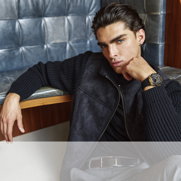 Man wearing a black jacket and watch sitting on a leather bench.