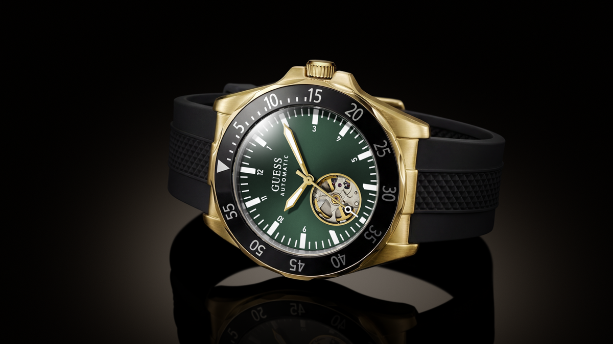 A Guess wristwatch with a gold-tone case, black bezel, dark green dial, and black textured strap, shown on a reflective black background.
