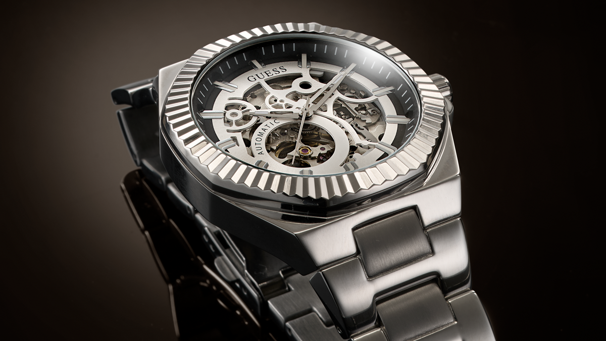 A close-up studio shot of a Guess skeleton wristwatch with a silver-tone metal case, black dial, and matching metal link bracelet against a dark gradient background.