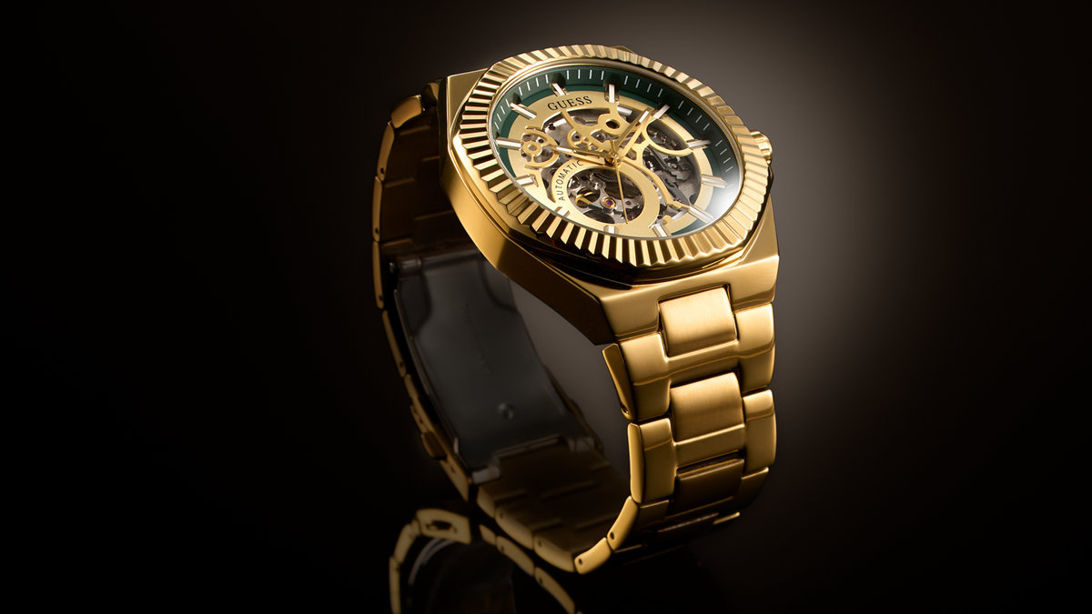 A luxury gold-tone skeleton watch with a matching gold-tone metal link bracelet, displayed on a reflective black surface against a dark background.