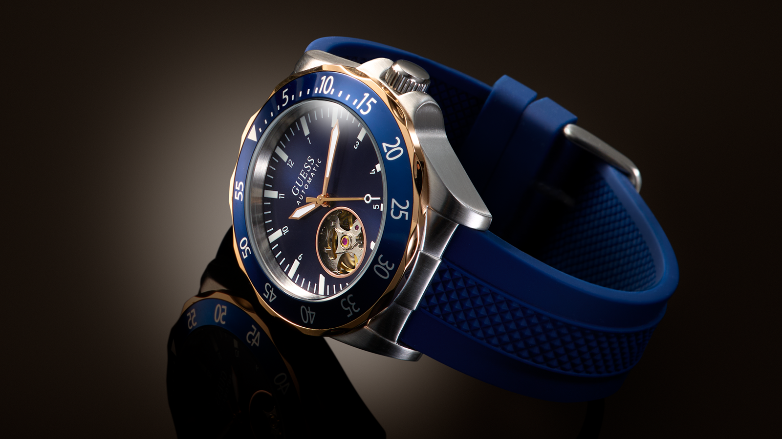 A GUESS wristwatch with a blue silicone strap, a blue dial, a blue rotating bezel, and a silver-toned case with a gold-tone inner ring. The watch is photographed at an angle against a dark, moody background.