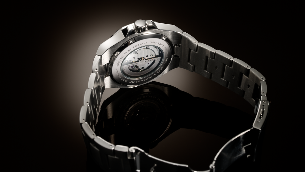 Close-up of a stainless steel watch with a transparent back case showing the internal mechanical movement, set against a dark reflective background.