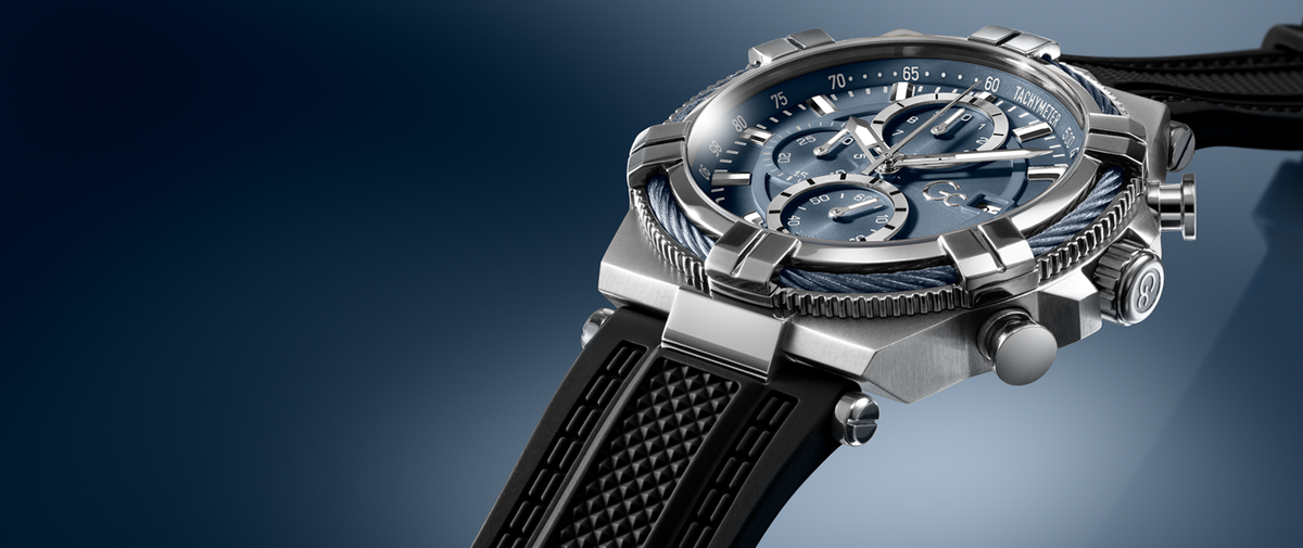 A luxury chronograph watch with a silver-toned titanium case, a blue dial with three subdials, and a black textured rubber strap, shown at an angle against a dark blue gradient background.
