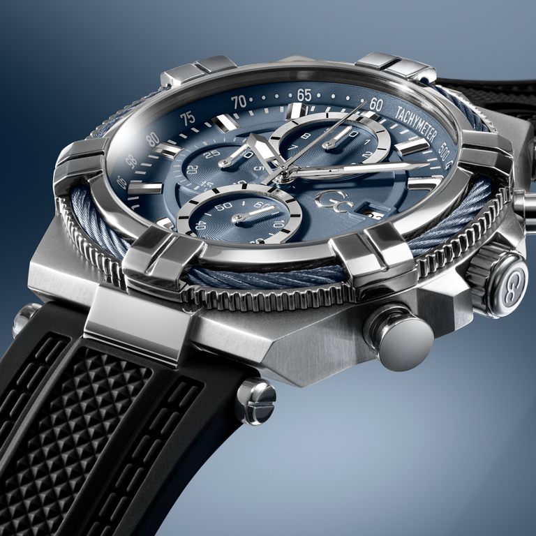 A close-up studio shot of a luxury chronograph watch with a brushed silver-tone case, a blue dial with three subdials, a blue rope detail around the bezel, and a black textured strap against a blue gradient background.