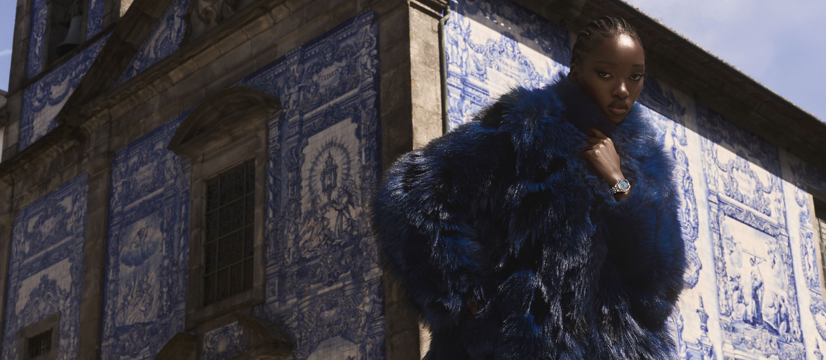 A model wearing a voluminous royal blue faux fur coat stands in front of a historic building featuring blue and white Portuguese azulejo tiles.