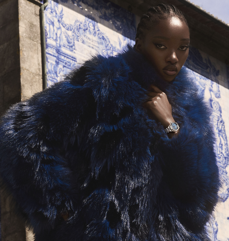 A model wearing a voluminous, luxurious deep blue faux fur coat, posing in front of a blue and white tiled wall.