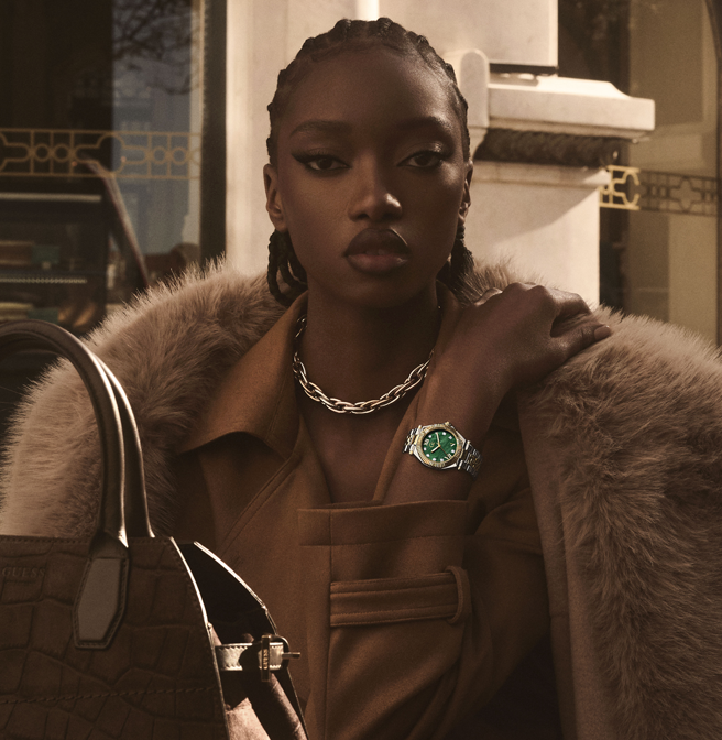 A model in a brown trench coat with a fur collar wears a green dial watch with a silver-tone case and chain necklace, holding a brown croc-embossed handbag in a warm, sunlit urban setting.