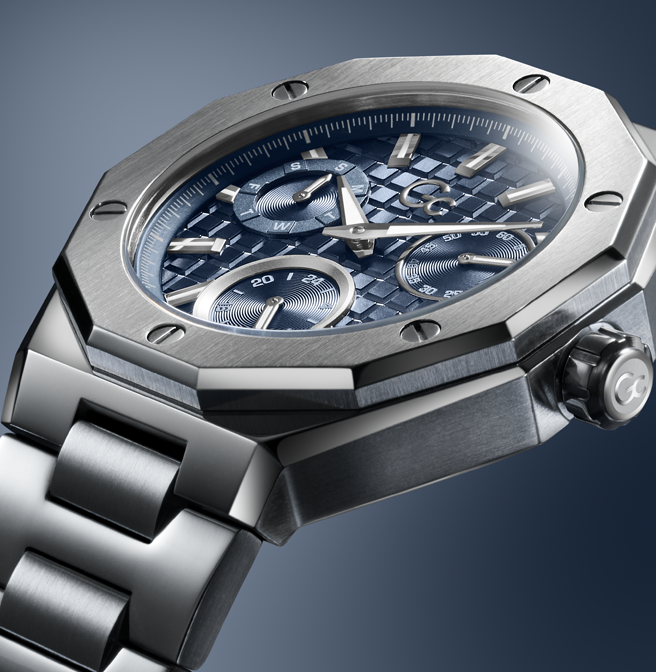 A close-up of a luxury watch with a brushed silver-tone octagonal case, a blue textured dial with three subdials, and a matching silver-tone metal link bracelet, set against a dark blue gradient background.