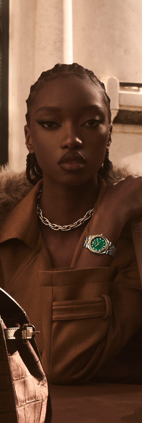 A close-up lifestyle shot of a model wearing a brown leather jacket with a fur collar, a silver chain necklace, and a wristwatch featuring a green dial and silver case.