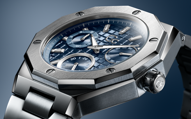 A close-up shot of a luxury watch with an octagonal stainless steel case, a textured blue dial with three subdials, and a matching blue and silver metal link bracelet, set against a dark blue gradient background.
