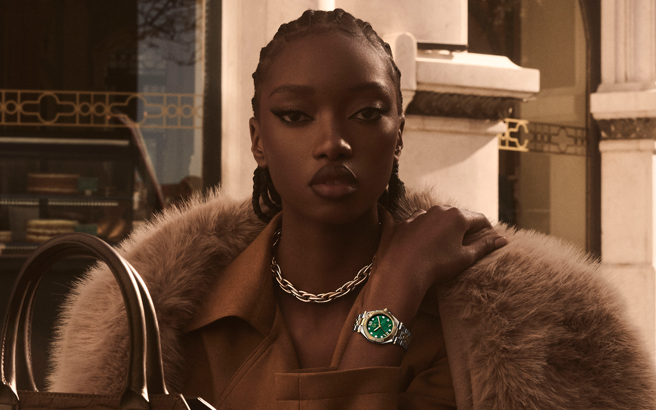 A model wearing a brown fur-trimmed jacket and a chunky silver chain necklace, wearing a gold watch with a green dial on her wrist, set against a warm-toned architectural background.