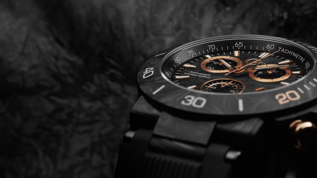 A close-up of a black chronograph watch with a black dial, white and orange numerals, and rose-gold-toned subdials and hands, set against a dark textured background.