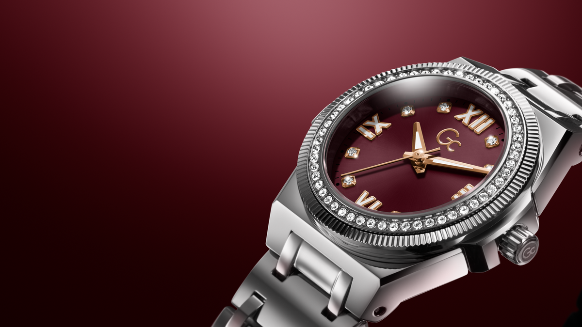 A close-up of a luxury watch with a silver stainless steel bracelet, a bezel set with small clear crystals, and a deep burgundy dial with gold-tone hands and Roman numeral hour markers.