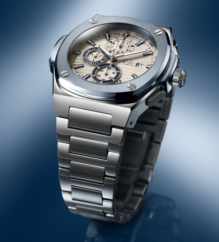 A luxury chronograph wristwatch with a brushed silver stainless steel case and integrated link bracelet, featuring a beige dial with black subdials and markers, set against a blue gradient background.