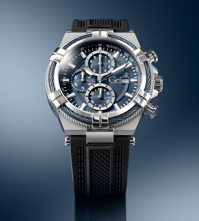 A G-CO brand chronograph watch with a silver stainless steel case, a dark blue dial with three subdials, and a black textured rubber strap, shown against a dark blue gradient background.