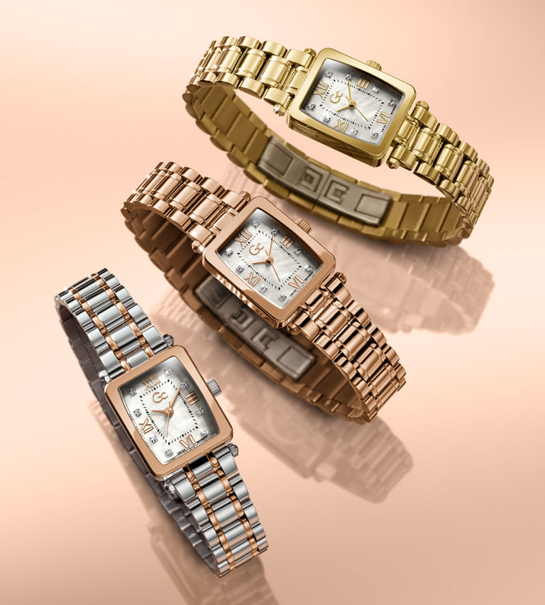 Three women's analog wristwatches with rectangular faces and linked metal bracelets are arranged on a soft peach background. One watch is gold-tone, one is rose gold-tone, and one is two-tone silver and gold-tone.