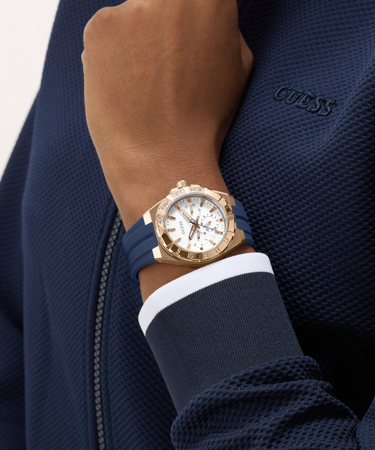 Gold watch on a wrist with a blue strap, wearing a navy jacket with 'Guess' branding.