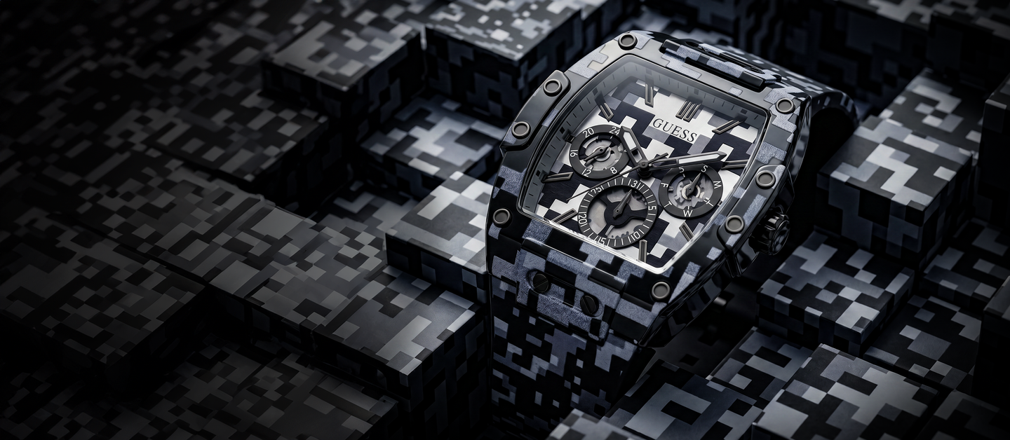 A GUESS chronograph watch with a black and white digital camouflage patterned case and strap, displayed on a dark geometric block background.