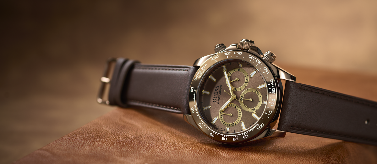 A GUESS chronograph watch with a dark brown leather strap, a bronze-toned case, and a dark dial with olive green subdials, resting on a textured brown leather surface.