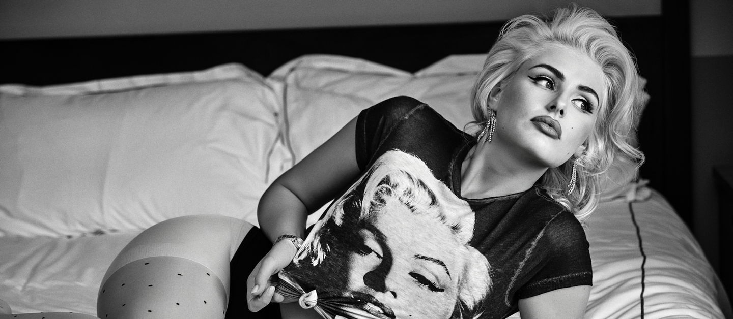 A black and white lifestyle photograph of a blonde woman reclining on a bed, wearing a black t-shirt with a large graphic print of Marilyn Monroe's face.