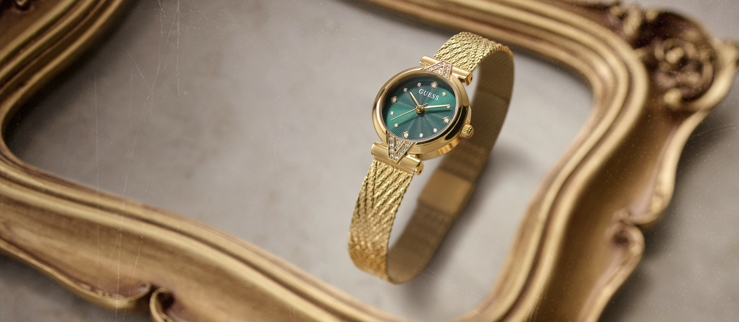 A gold-tone mesh bracelet watch with a dark teal-green dial, resting on an ornate gold picture frame against a neutral gray background.