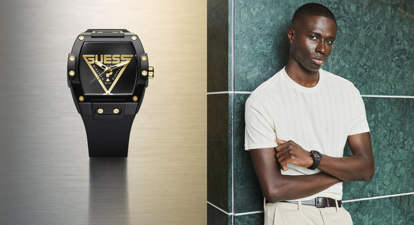Black Guess watch with gold accents on a metallic background, next to a man wearing a white shirt and beige pants against a green tiled wall.