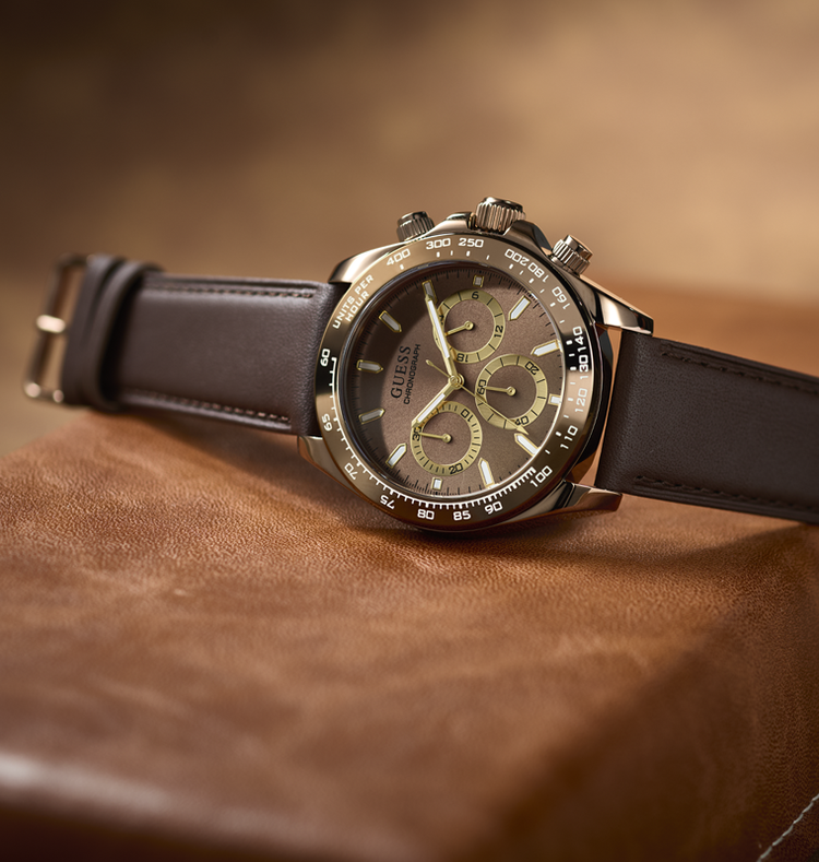 A Guess men's chronograph watch with a dark brown leather strap, a brushed stainless steel case, and a dark brown dial with olive-green subdials, resting on a textured brown leather surface.