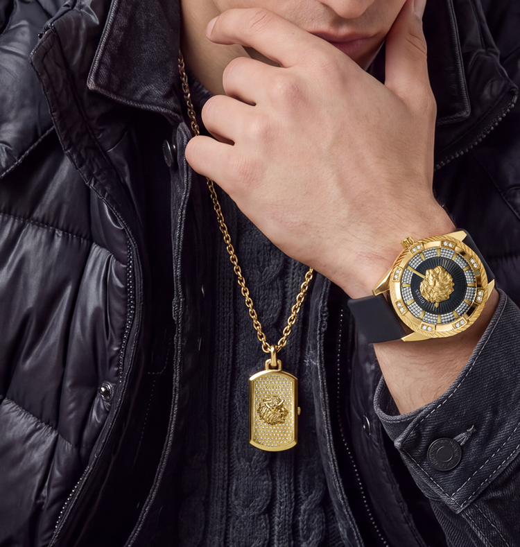A close-up lifestyle shot of a person wearing a gold Versace pendant necklace and a gold Versace wristwatch with a black dial and rhinestone bezel, styled with a black puffer jacket.