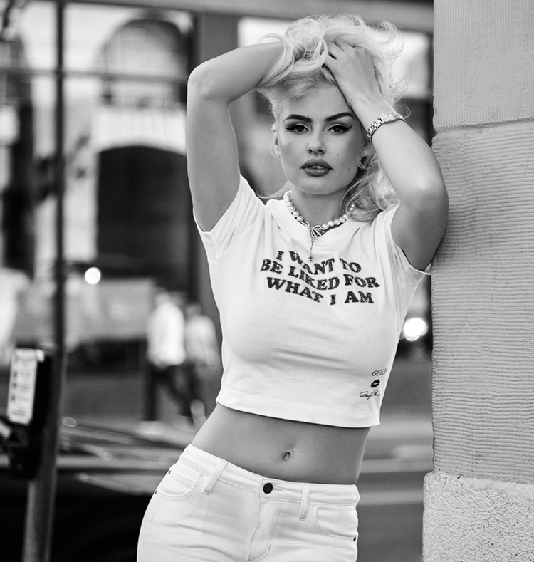 A black and white lifestyle photograph of a woman posing against a textured wall, wearing a white graphic crop top with the text 'I WANT TO BE LIKED FOR WHAT I AM' and white jeans.