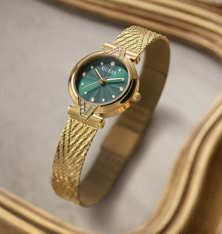A gold-tone Guess wristwatch with a textured mesh band and a green sunburst dial, resting on a reflective gold surface.