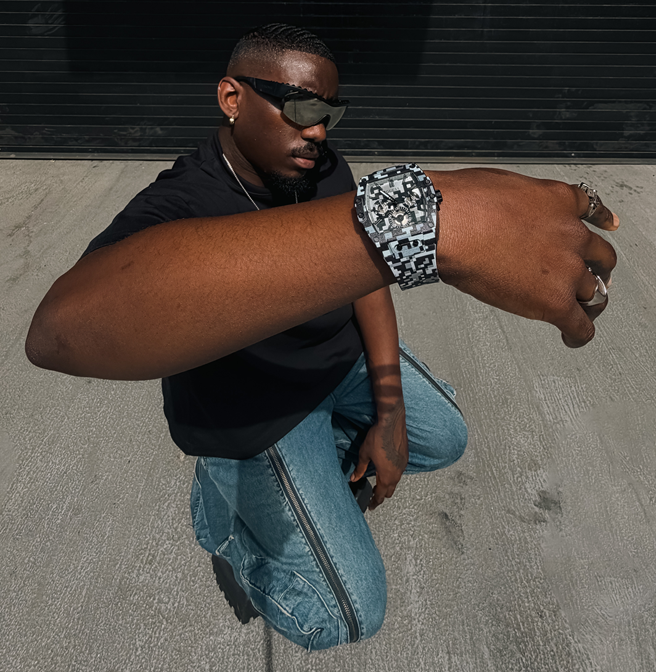 A man wearing a chunky silver-toned watch with a black and white camouflage patterned band, posing outdoors on a concrete surface in front of a dark garage door.