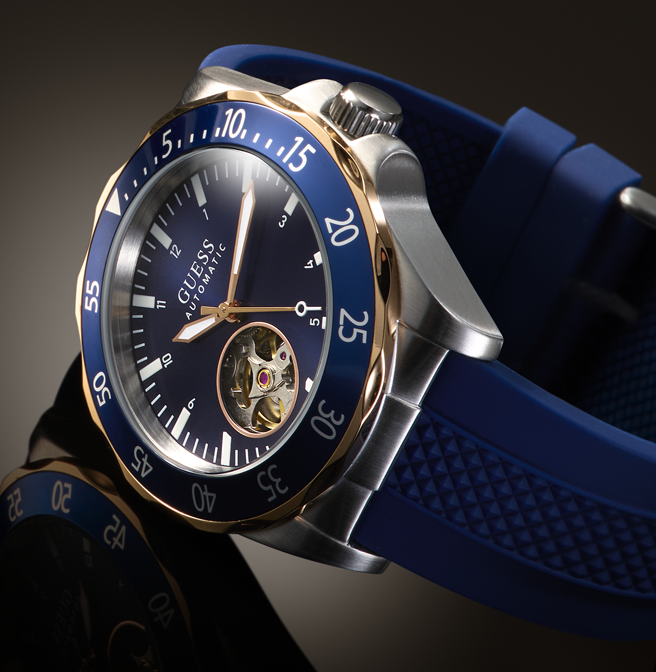 A Guess Automatic watch with a blue dial, blue silicone strap, gold-tone bezel accents, and a silver-tone stainless steel case, shown in a close-up studio shot.