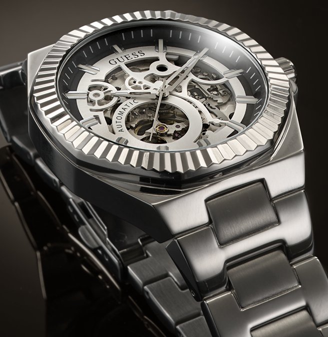 A close-up of a Guess skeleton wristwatch with a dark gray and silver-tone metal case, transparent dial showing internal gears, and a matching metal link bracelet.