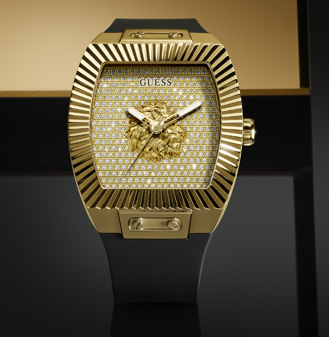 A luxury Guess wristwatch with a gold-tone case, a diamond-embellished dial, a lion head motif at the center, and a black strap, shown against a dark and gold background.