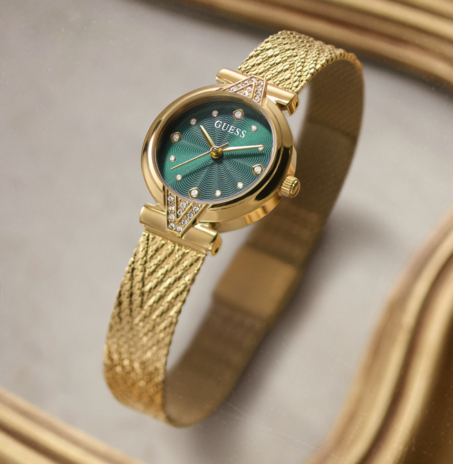 A gold-tone Guess wristwatch with a textured mesh band and a green dial, shown on a neutral gray background with soft gold bokeh.