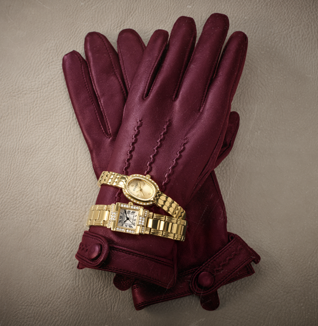 A pair of burgundy leather gloves with decorative stitching and cuff details, styled with two gold-tone wristwatches on a textured gray background.