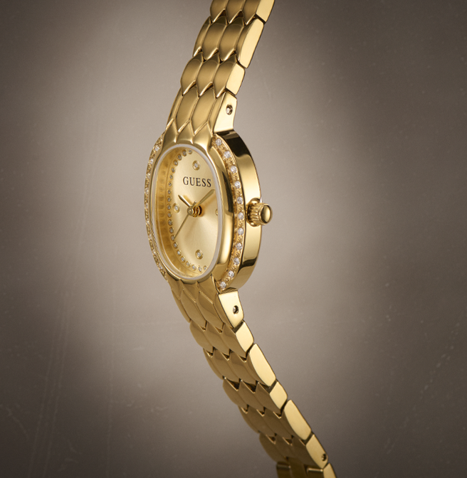 Gold-tone Guess watch with a diamond-accented bezel and matching gold-tone link bracelet, shown on a dark gray gradient background.