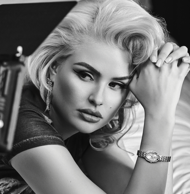 A black and white close-up portrait of a woman with platinum blonde hair, wearing a bracelet watch on her wrist and dangling earrings, resting her head on her hand.