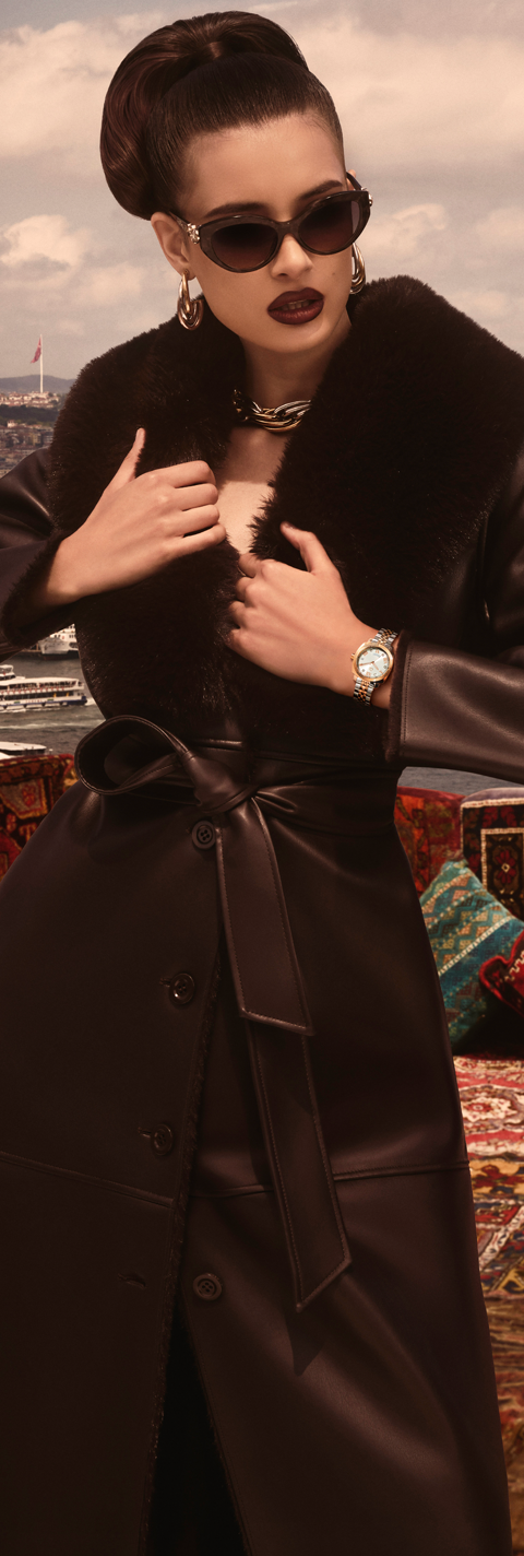 Woman in a dark coat and sunglasses adjusting her collar with a cityscape in the background