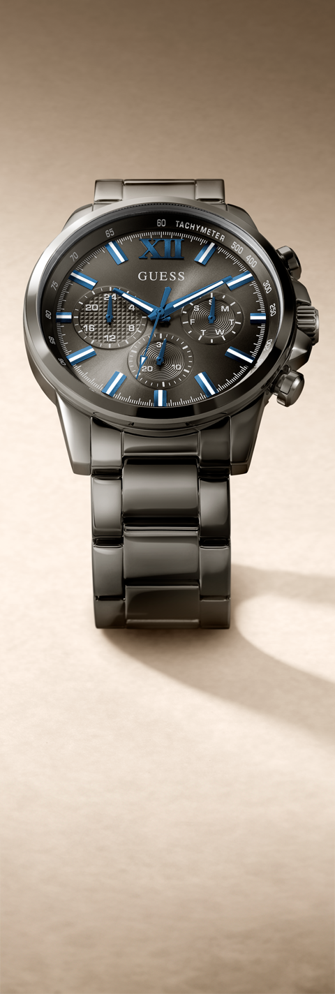 A Guess men's chronograph watch with a dark gray and black metallic case and bracelet, featuring a textured dark gray dial with blue luminous hands and markers, set against a warm beige background.