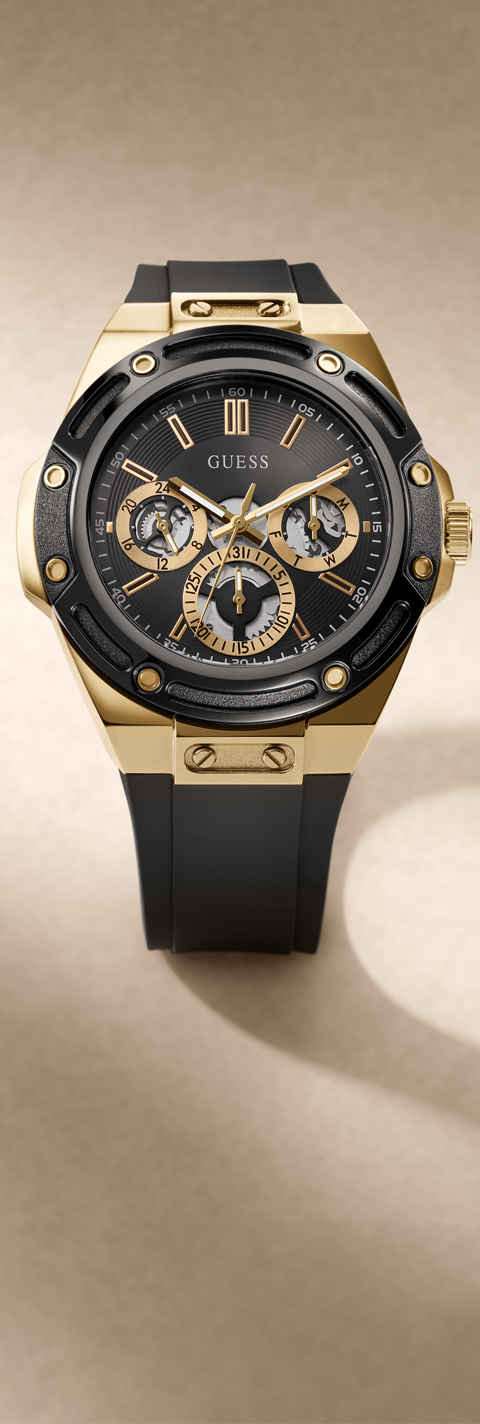A luxury wristwatch by Guess with a black rubber strap, a gold-tone case, and a black dial featuring three sub-dials and gold accents, set against a warm beige background.