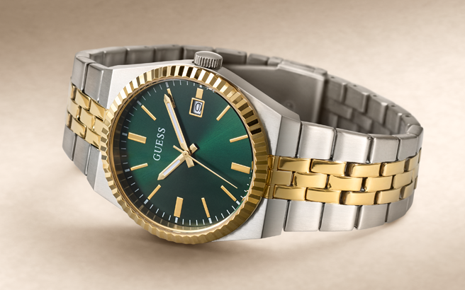 A Guess men's two-tone watch with a green dial, gold-tone bezel and indices, and a silver and gold-tone stainless steel link bracelet, shown on a neutral beige background.